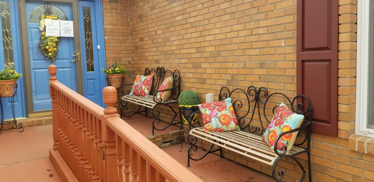 A cozy patio with wrought iron benches adorned with colorful pillows, set against a brick wall, featuring potted plants for decoration.
