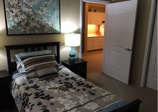 A cozy bedroom with a bed adorned with patterned bedding, a lamp on the bedside table, and an open door leading to a lit hallway.