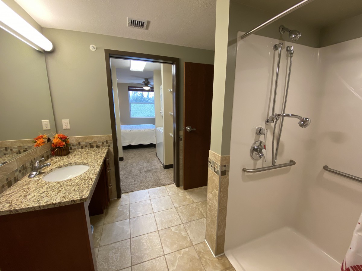 A view of a bathroom featuring a walk-in shower and sink with a mirror, leading into an adjacent room.