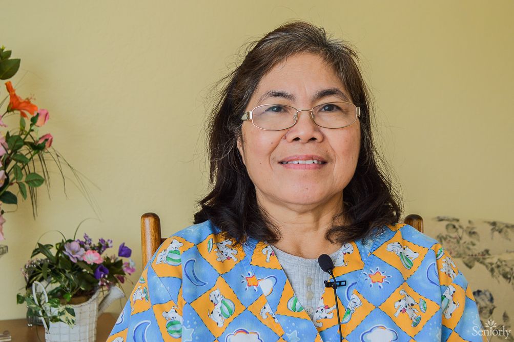 A smiling woman wearing glasses sits indoors with a floral arrangement in the background.