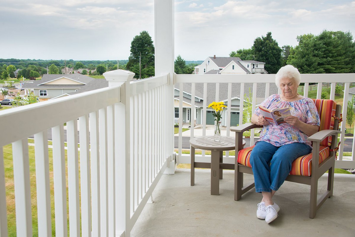 An elderly woman relaxes on a porch, enjoying a book with a view of a residential neighborhood.