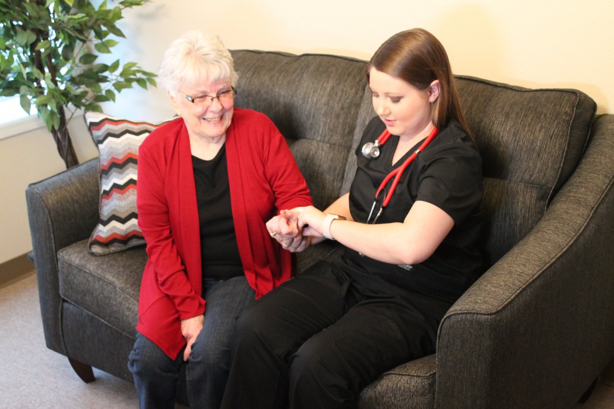 A healthcare professional attends to an elderly woman during a home visit, checking her wrist and providing support.
