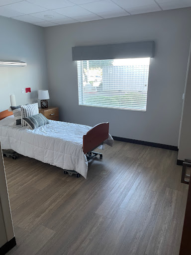 A bright and minimalistic hospital room featuring a single bed, bedside table, and large window.