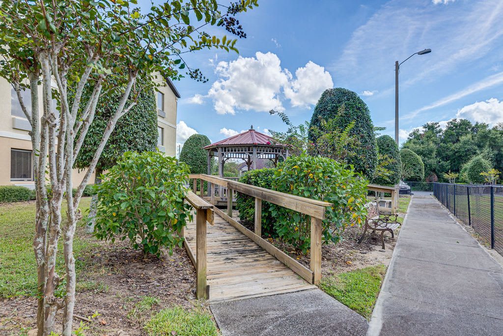 A serene garden scene featuring a wooden bridge leading to a gazebo, surrounded by trees and bushes under a partly cloudy sky.