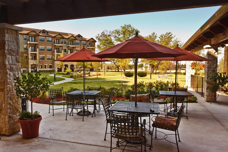 A serene outdoor patio area with tables and umbrellas overlooking a lush garden and building in the background.