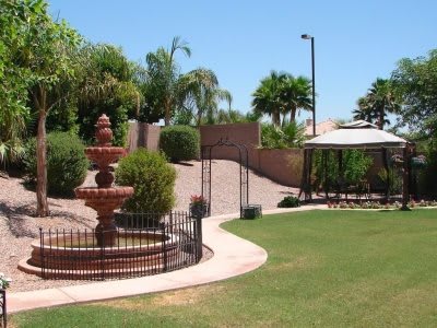 A landscaped garden featuring a fountain, gazebo, and paved pathway surrounded by trees and grass.