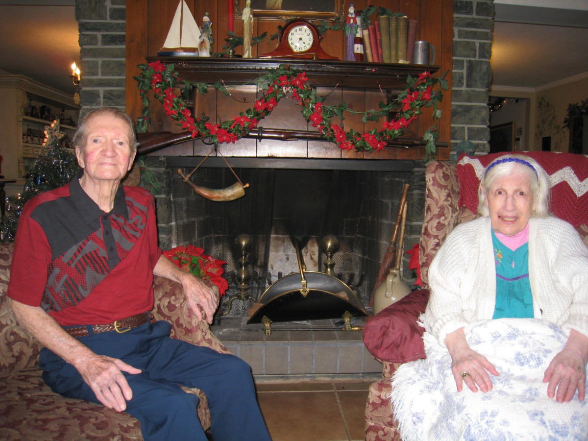 An elderly couple sitting in a cozy living room by the fireplace, adorned with holiday decorations.
