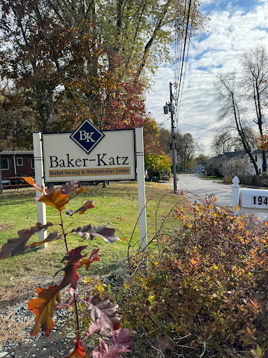 A welcoming sign for Baker-Katz along a scenic road surrounded by autumn foliage.
