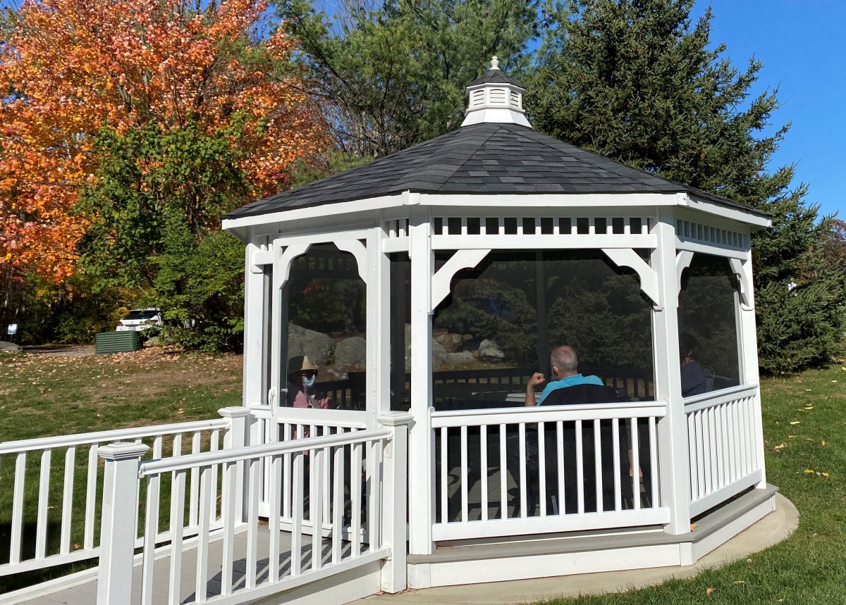 A person sits inside a white gazebo surrounded by trees with autumn foliage.