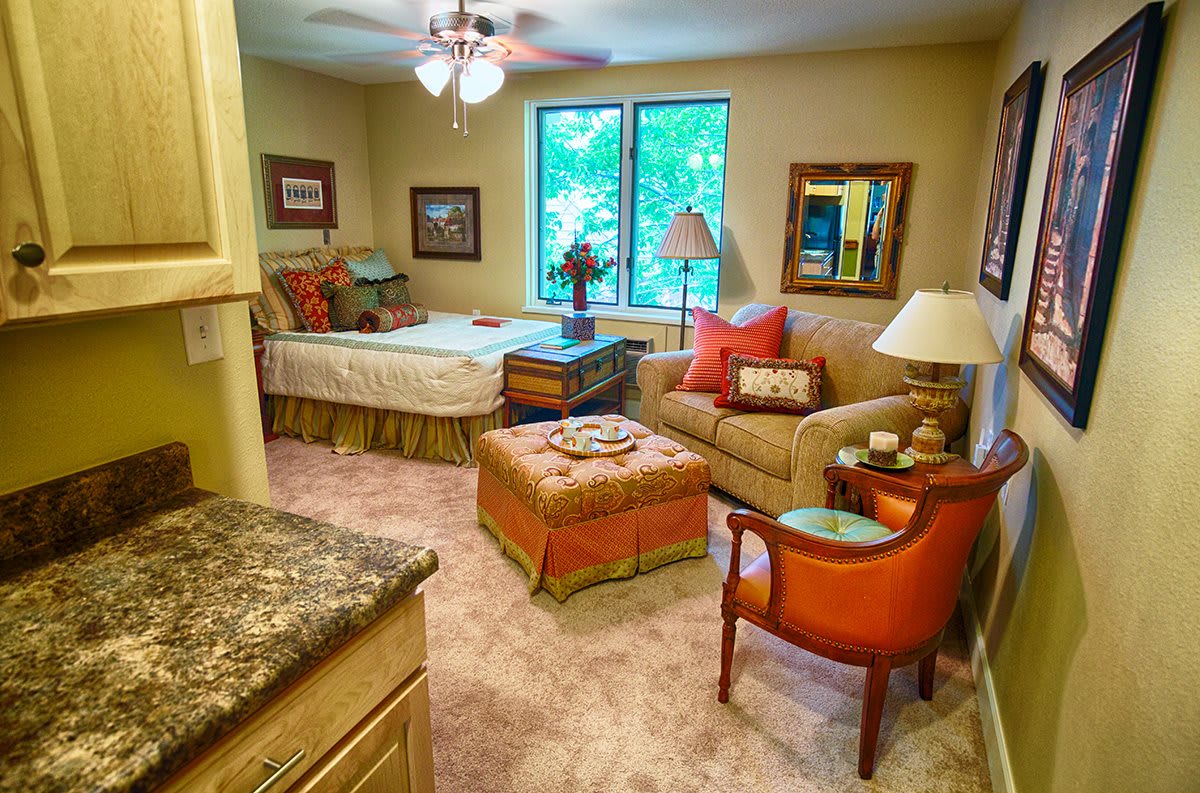 A cozy bedroom with a connected living area featuring a bed, sofa, and armchair adorned with colorful pillows. The room is warmly lit and includes large windows and tasteful decor.