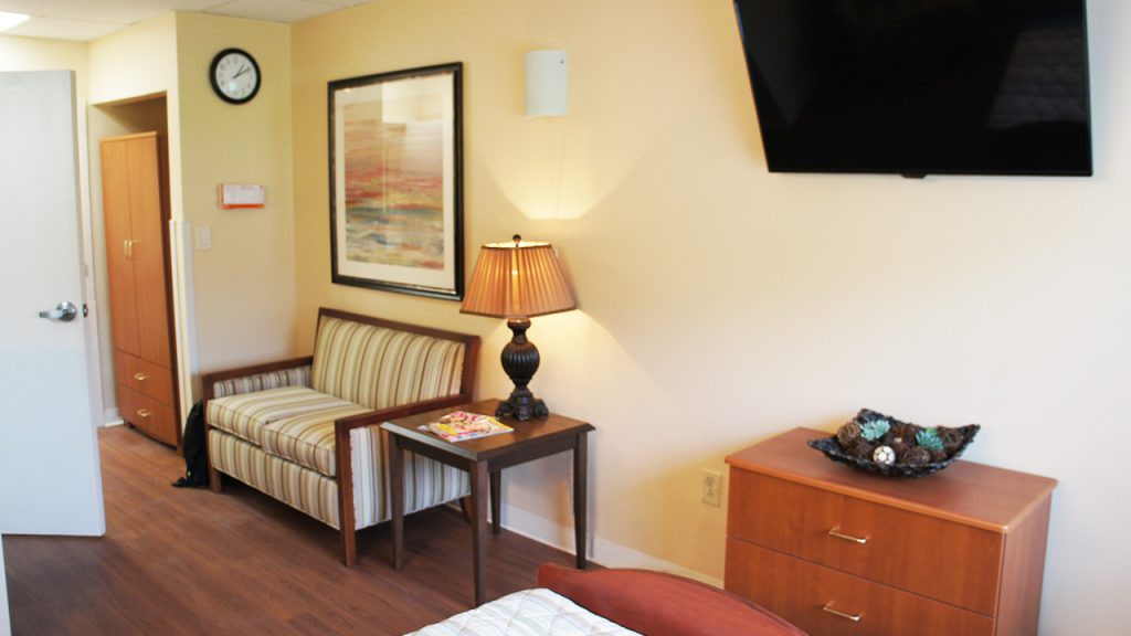 A cozy hotel room interior featuring a striped sofa, a side table with a lamp, a wall-mounted television, and artwork on the wall.