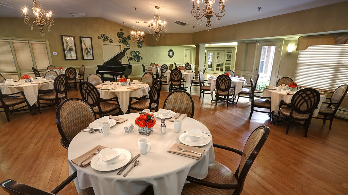 An elegant dining room with beautifully set tables, a grand piano, and charming chandeliers.