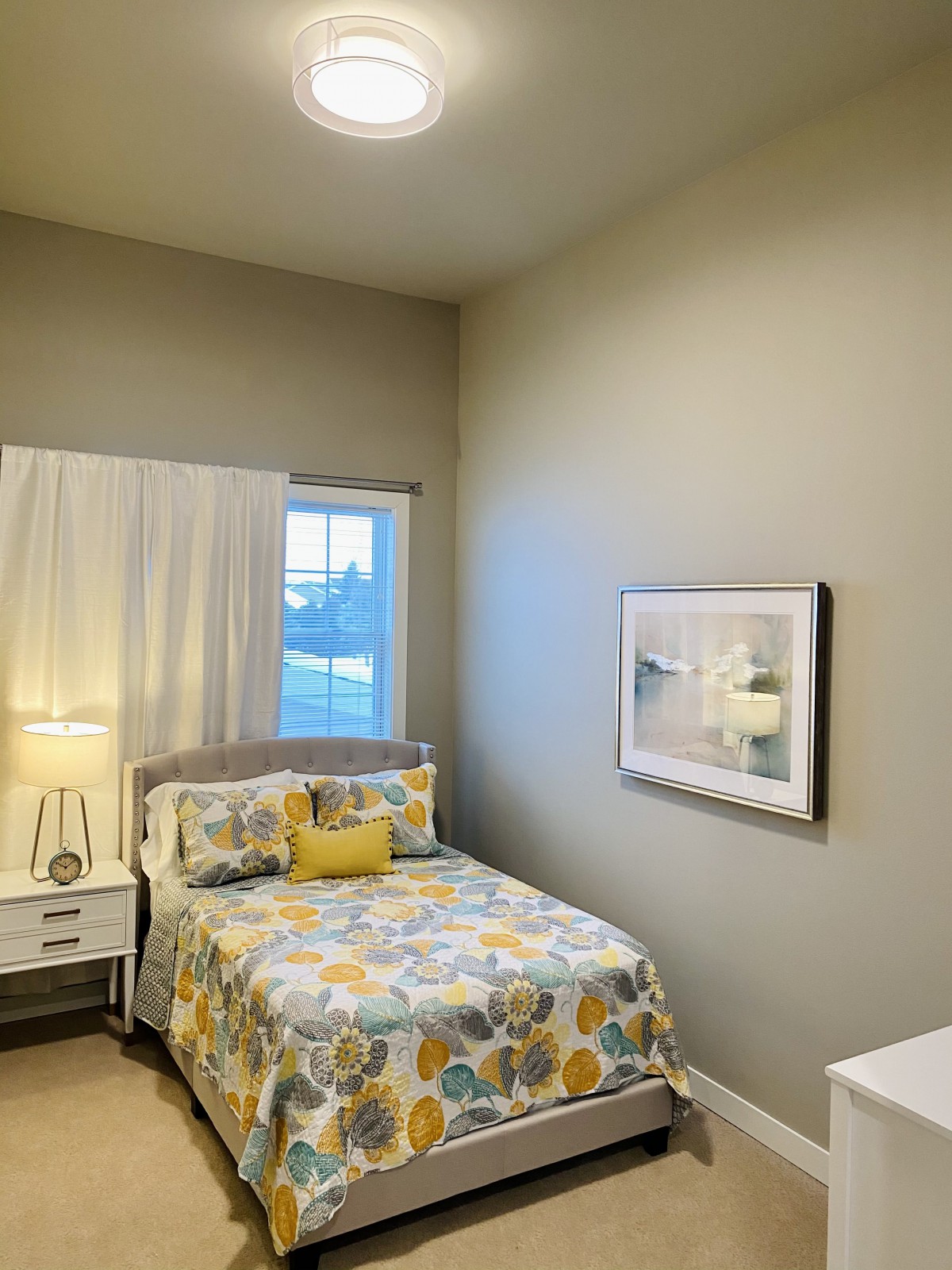 A cozy bedroom with a floral bedspread, bedside table with a lamp, and a picture on the wall.