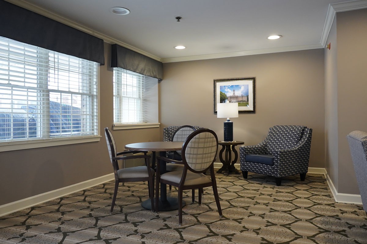 A cozy living room area with a round table, chairs, an armchair, and a lamp beside framed artwork.