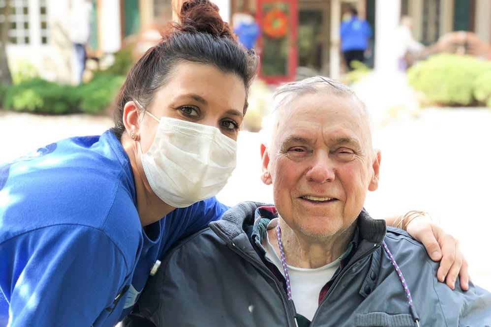 A nurse wearing a mask poses with an elderly patient outdoors.