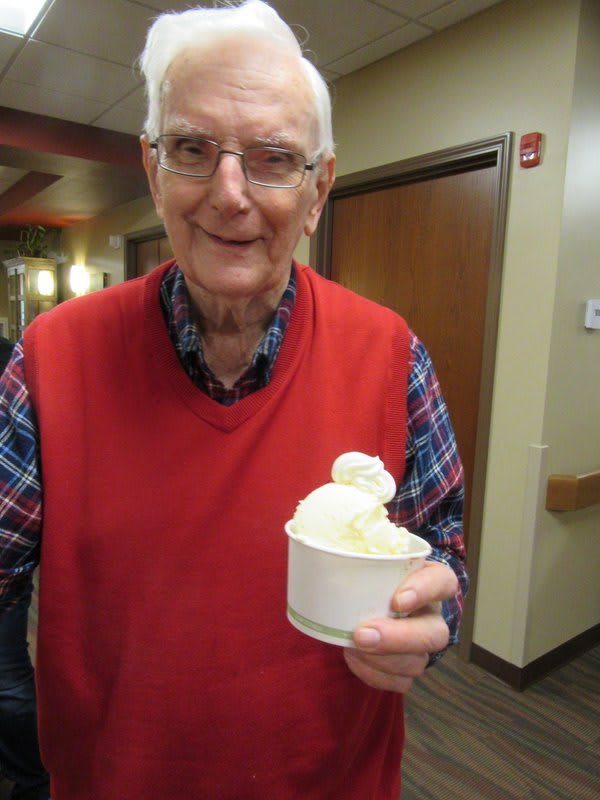 An elderly man in a red vest holding a cup of ice cream and smiling indoors.