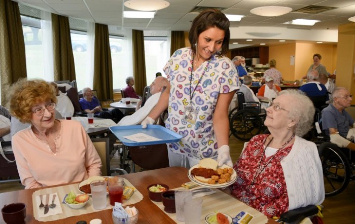 A caregiver serves a meal to residents in a nursing home dining area.