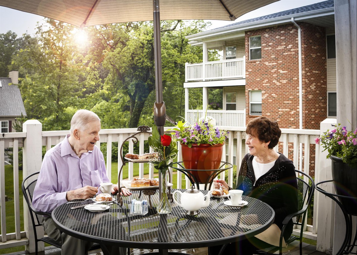 An elderly couple enjoys tea on a sunny patio, surrounded by greenery.