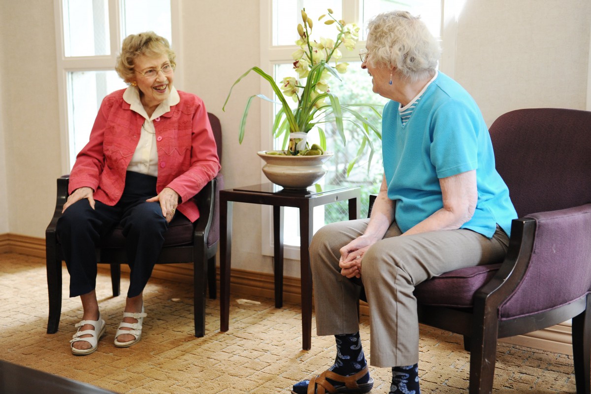 Two elderly women engage in a conversation while seated indoors.