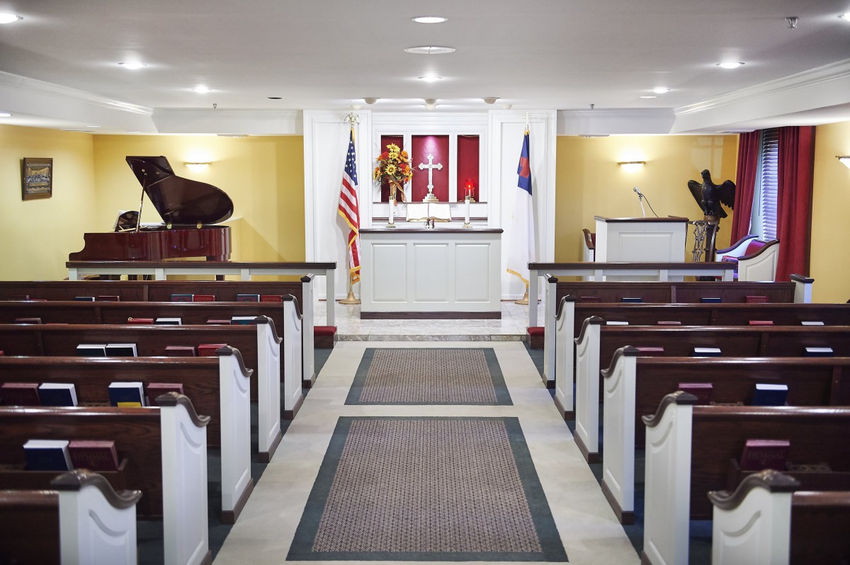 A small chapel interior with pews facing an altar, flanked by pianos and flags.