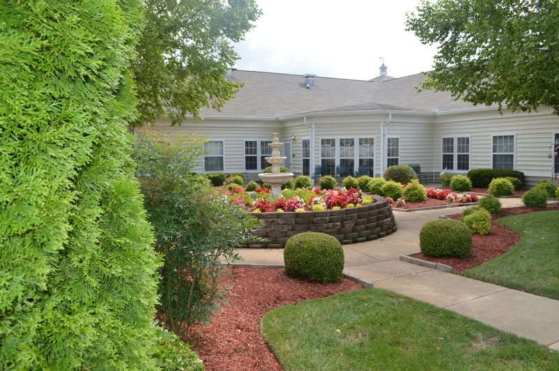 A beautifully landscaped garden with a central fountain surrounded by colorful flowers and trimmed bushes, leading to a residential building.