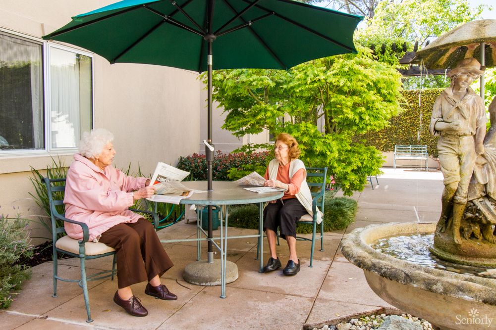 Two people sitting at a patio table under an umbrella, reading newspapers in a garden setting.