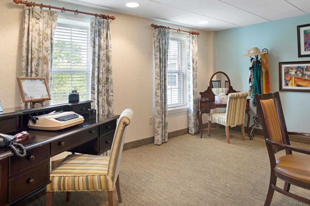 A cozy room with a desk and vanity setup, featuring elegant chairs and ample natural light through large windows.