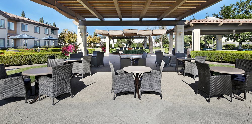 An outdoor patio area featuring wicker tables and chairs, set under a shaded pergola with a garden in the background.