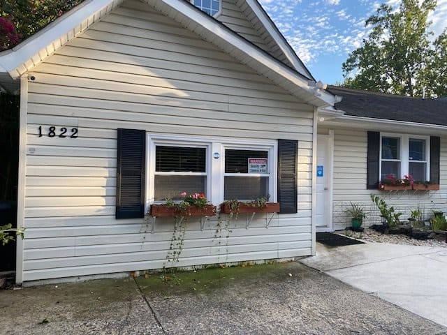 A small house with white siding and blue shutters, featuring a front garden and paved walkway.