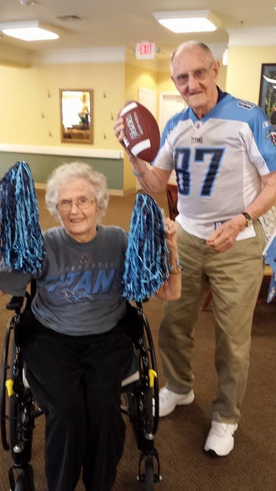 An elderly person in a wheelchair holds blue pom poms while another person stands nearby holding a football.