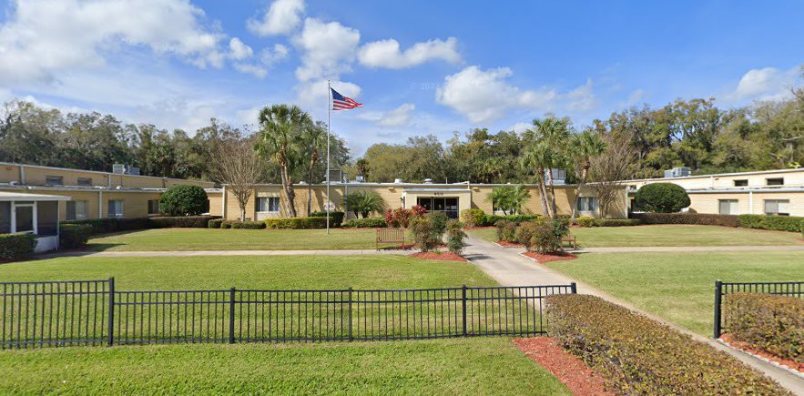 A single-story building with an American flag on its roof, surrounded by a well-maintained lawn, palm trees, and a black fence under a partly cloudy sky.