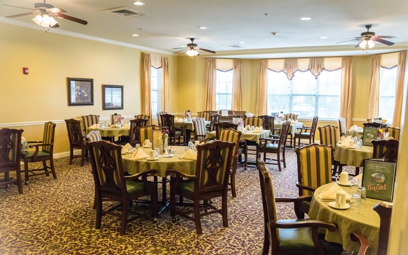 A spacious dining room with neatly arranged tables and chairs, featuring large windows and warm decor.