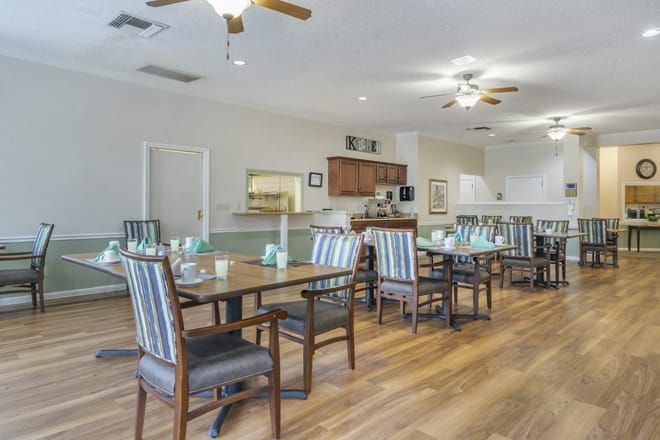 A spacious dining area with striped chairs and wooden tables, featuring ceiling fans and a kitchen in the background.