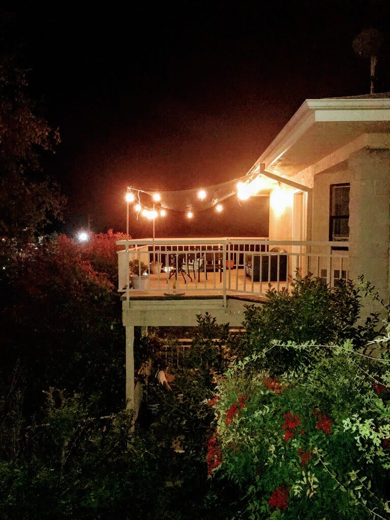 A cozy outdoor deck illuminated by string lights at night, surrounded by lush garden greenery.