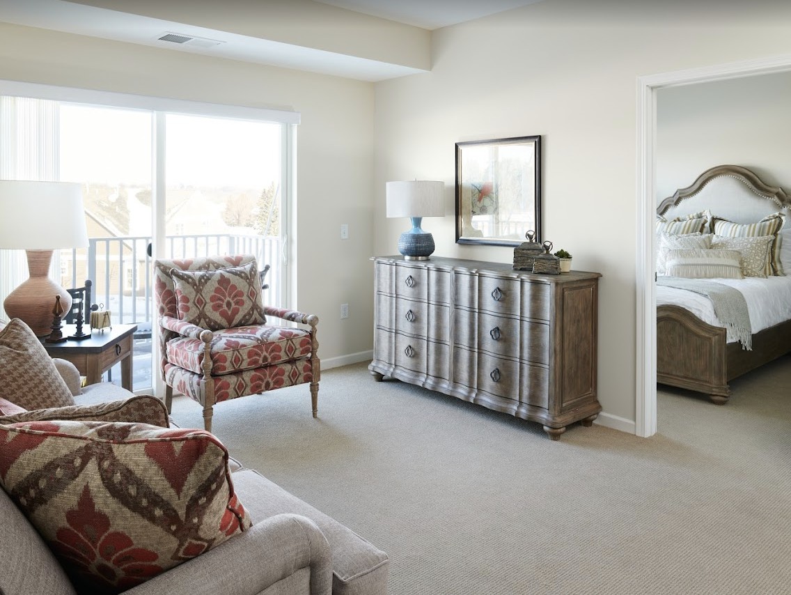 A cozy living room featuring patterned armchairs, a rustic sideboard, and large windows letting in natural light.