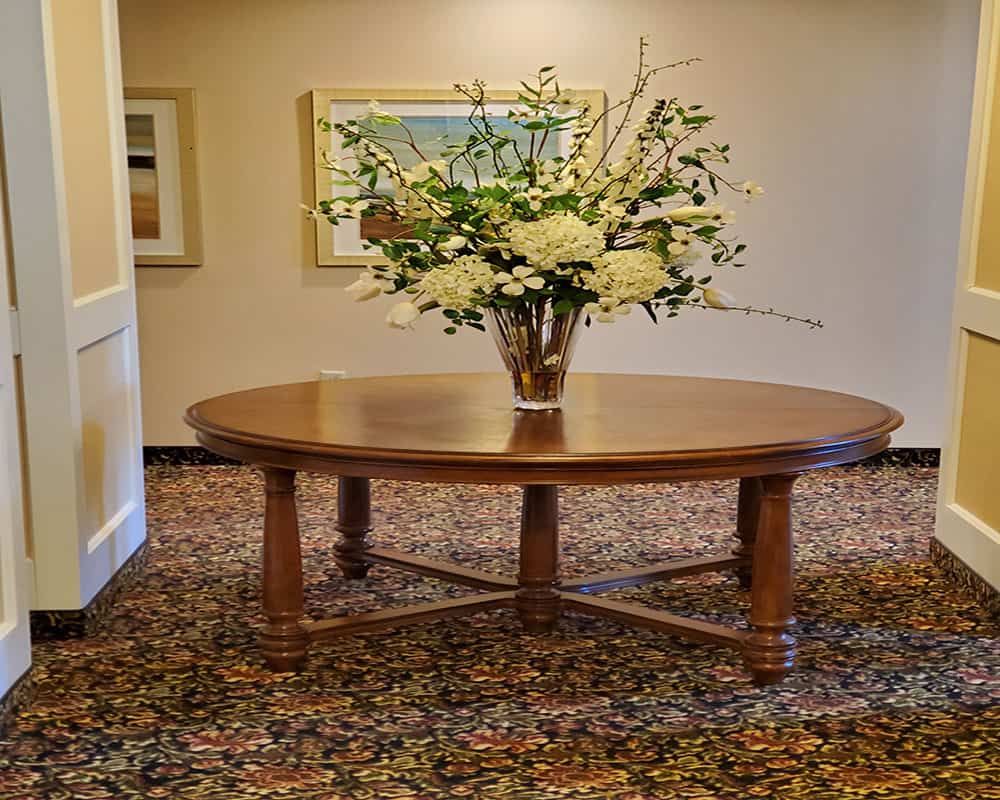 A round wooden table adorned with a vase of white and green flowers, set in a well-decorated interior space with patterned carpet and framed artwork on the walls.