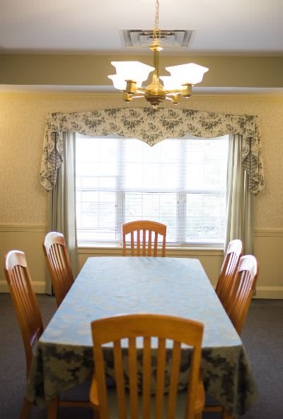 A dining room with a wooden table and six chairs, featuring a large window with patterned curtains and a chandelier overhead.