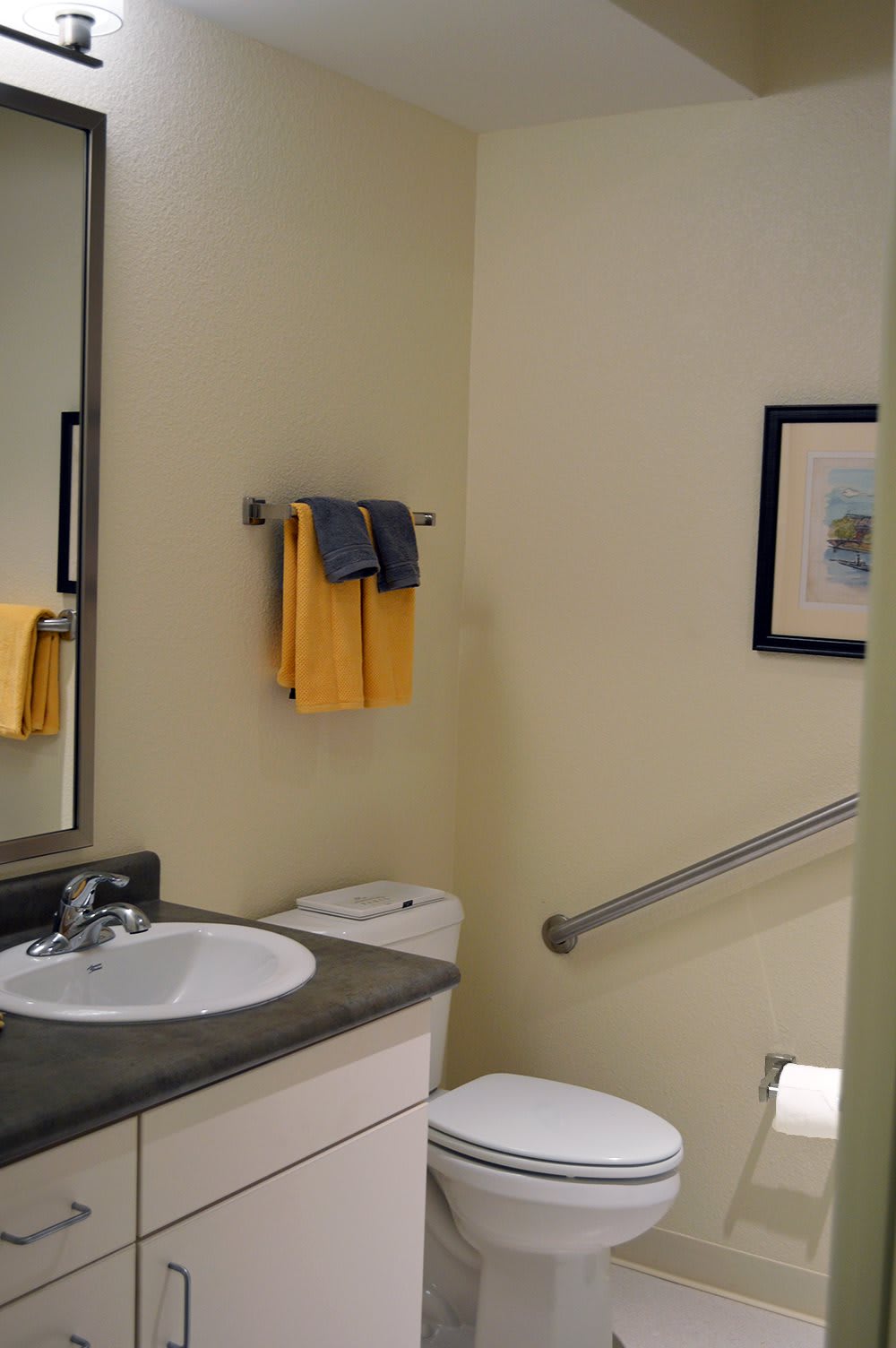 A neatly organized bathroom with a sink, mirror, yellow towels, and a handrail next to the toilet.
