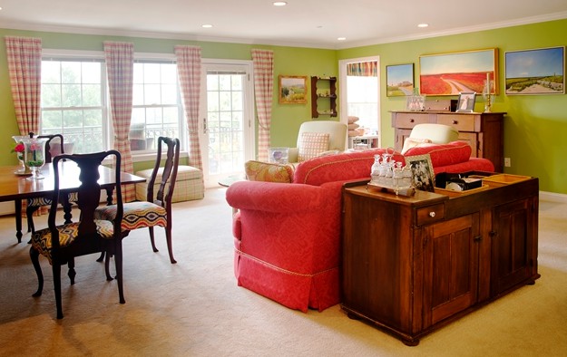 A cozy living room featuring a red sofa, chairs, and a dining table by large windows with checkered curtains.