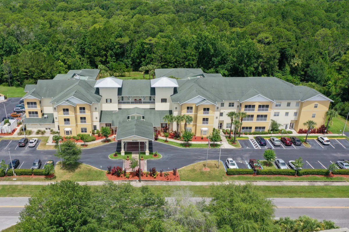 Aerial view of a residential building surrounded by lush greenery with a parking lot in front.