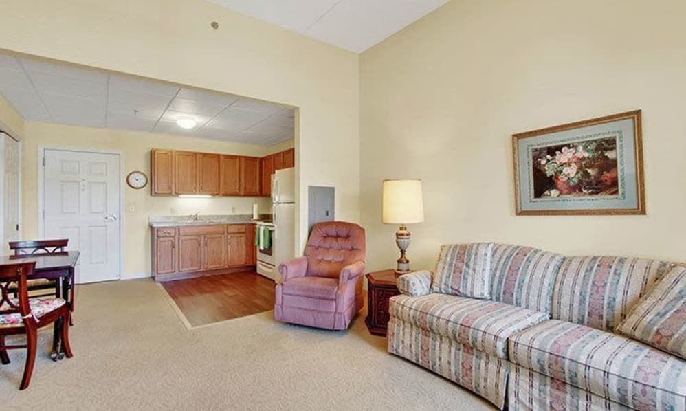 A cozy living room area with a view of the kitchen, featuring a comfortable sofa, recliner chair, and a floor lamp.