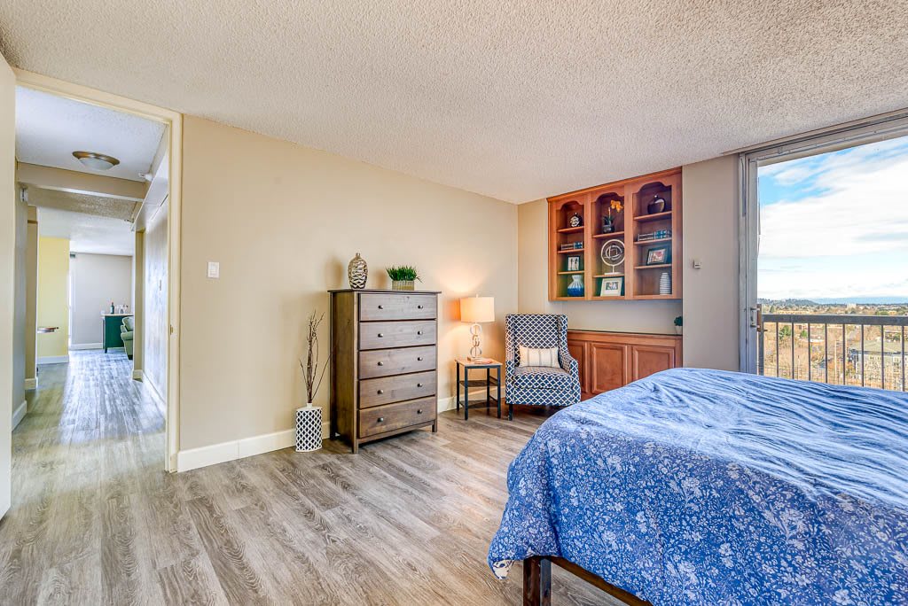 A cozy bedroom featuring wood flooring, a blue bedspread, a tall dresser, and a comfortable chair with a lamp for reading.