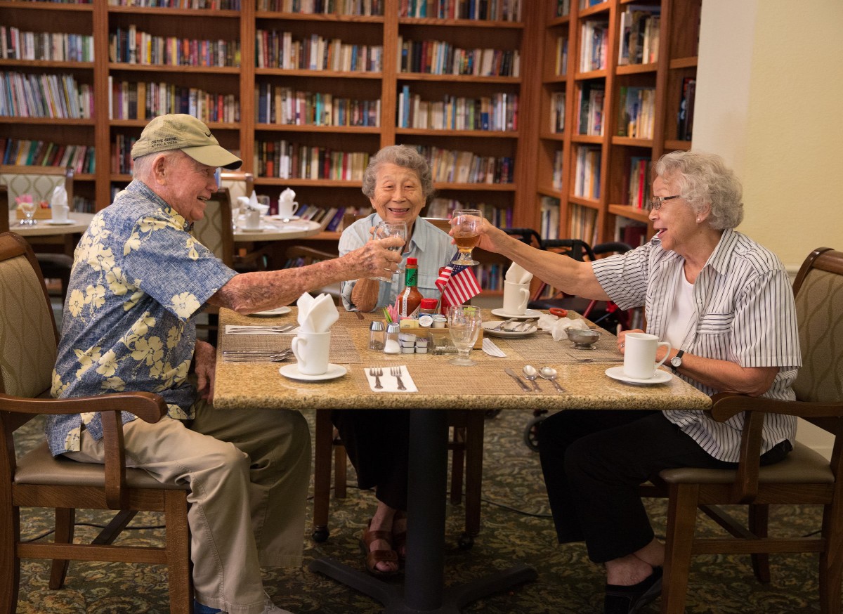 Three elderly individuals enjoy a meal together in a cozy library setting, raising their glasses in a toast.