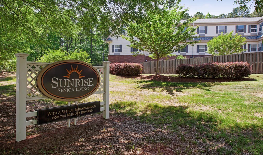 A sunny day view of a building, with a 'Sunrise Senior Living' sign in the foreground, surrounded by trees and well-maintained landscaping.