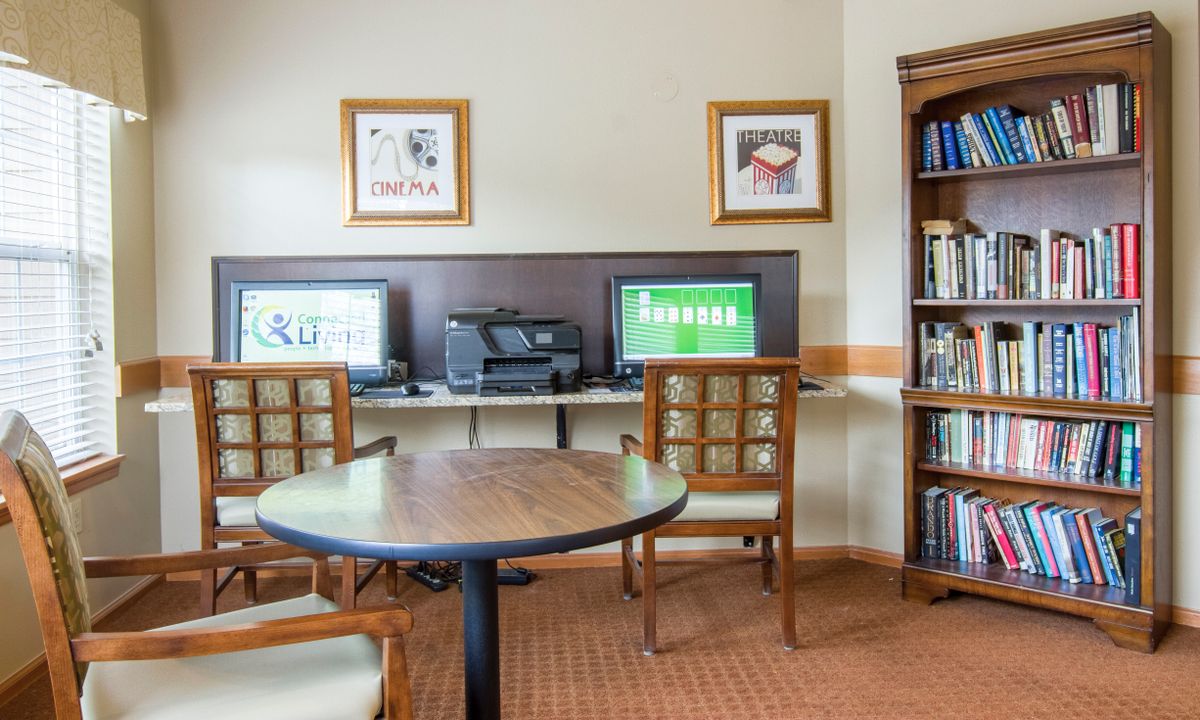 A small library or computer room with tables, chairs, and computers alongside bookshelves.