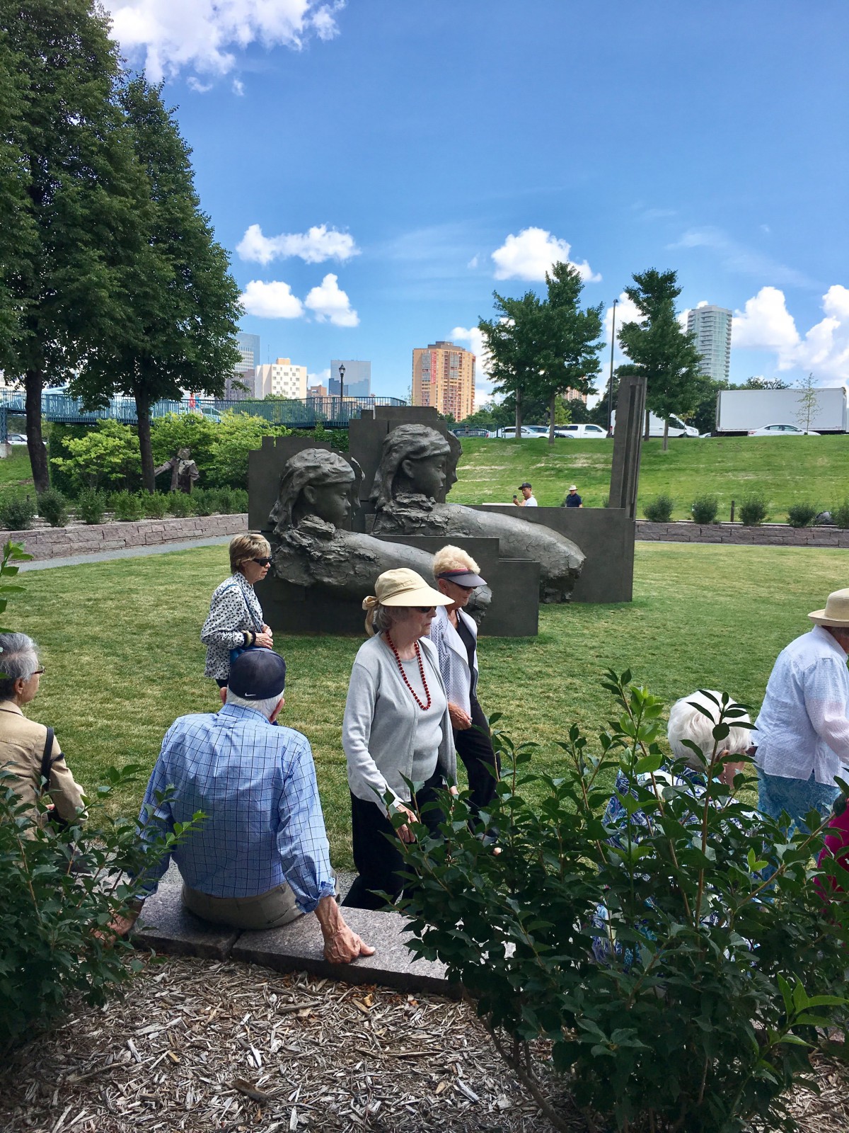 A group of people enjoying a sunny day in a park with a large sculpture and city skyline in the background.