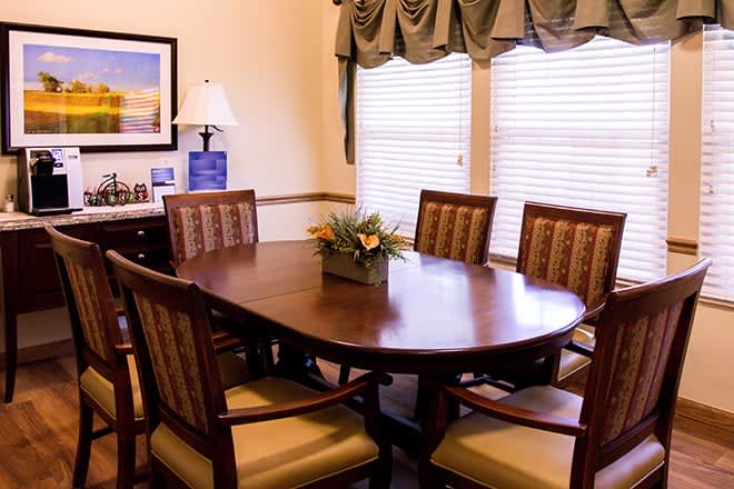 A cozy dining room with a wooden table, surrounded by chairs, and adorned with a floral centerpiece. The room features large windows with curtains, a side table with a lamp, and wall art.