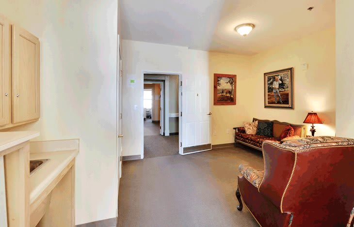 A cozy living room with a sofa and armchair, adorned with paintings on the walls, leading to an open hallway.
