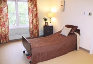 A cozy bedroom featuring a single bed with a brown cover, a nightstand with a lamp, and floral curtains by the window.