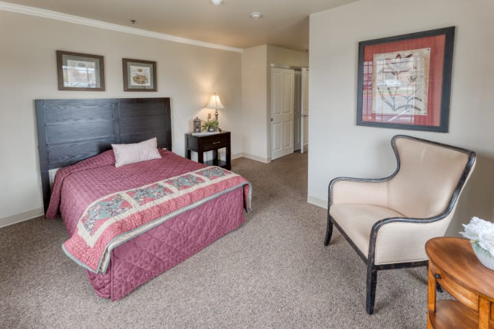 A cozy bedroom with a pink bedspread, featuring a chair and a nightstand with a lamp.